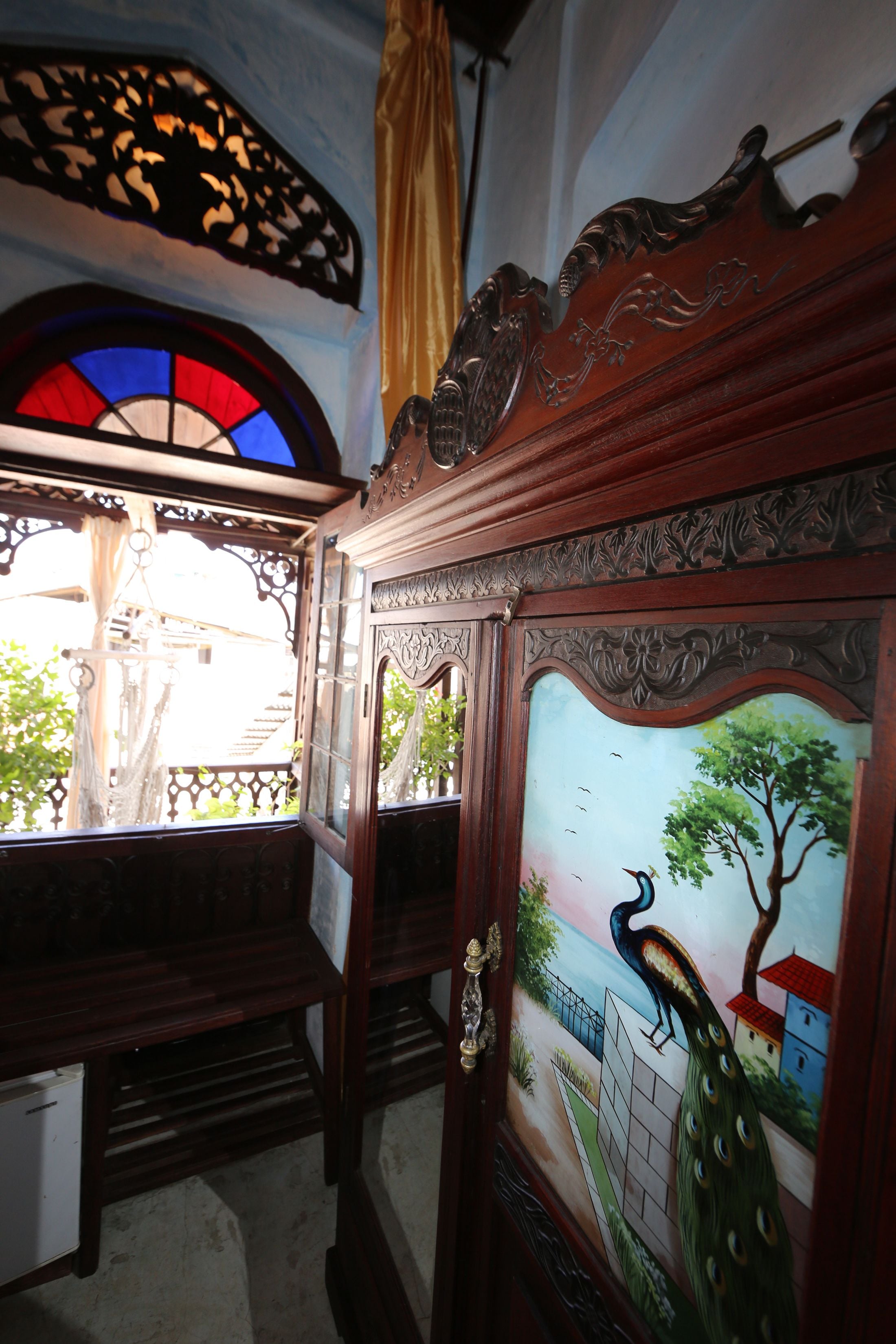 A room with a wooden door with intricate carvings, a stained glass window, and a mural of a peacock on the wall.
