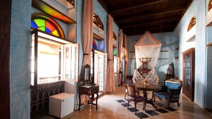 A room with a large window, a wooden cabinet, a table with chairs, and a hanging canopy. - Emerson Spice Hotel (Zanzibar Town)