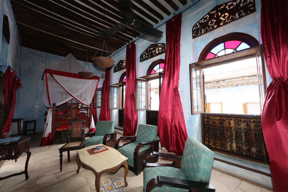 A room with a traditional design, featuring a bed with a canopy, several chairs, a table, and a large window with red curtains.