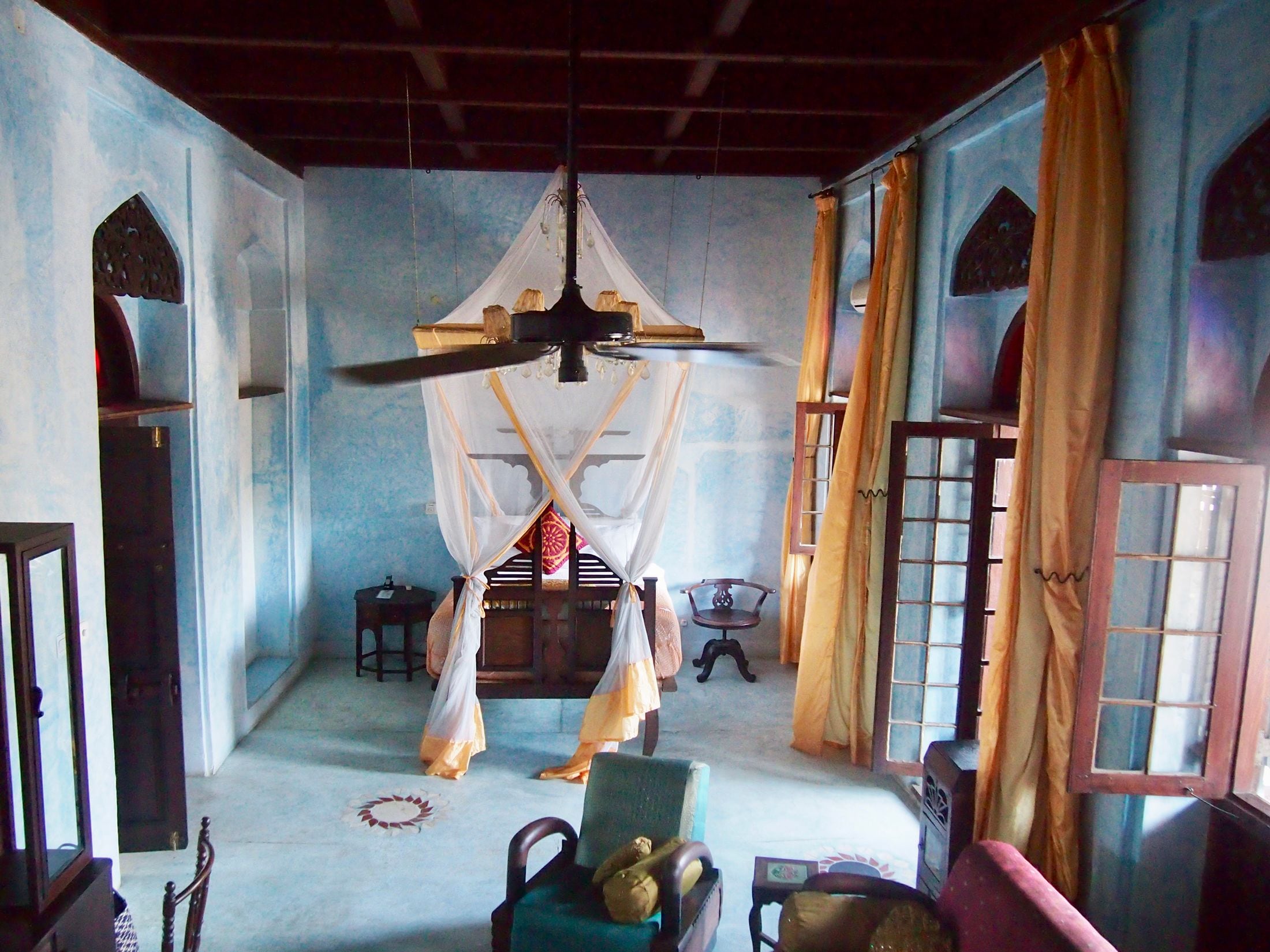 Una habitación con cama bajo un dosel, una silla, una mesita y una ventana con cortinas.