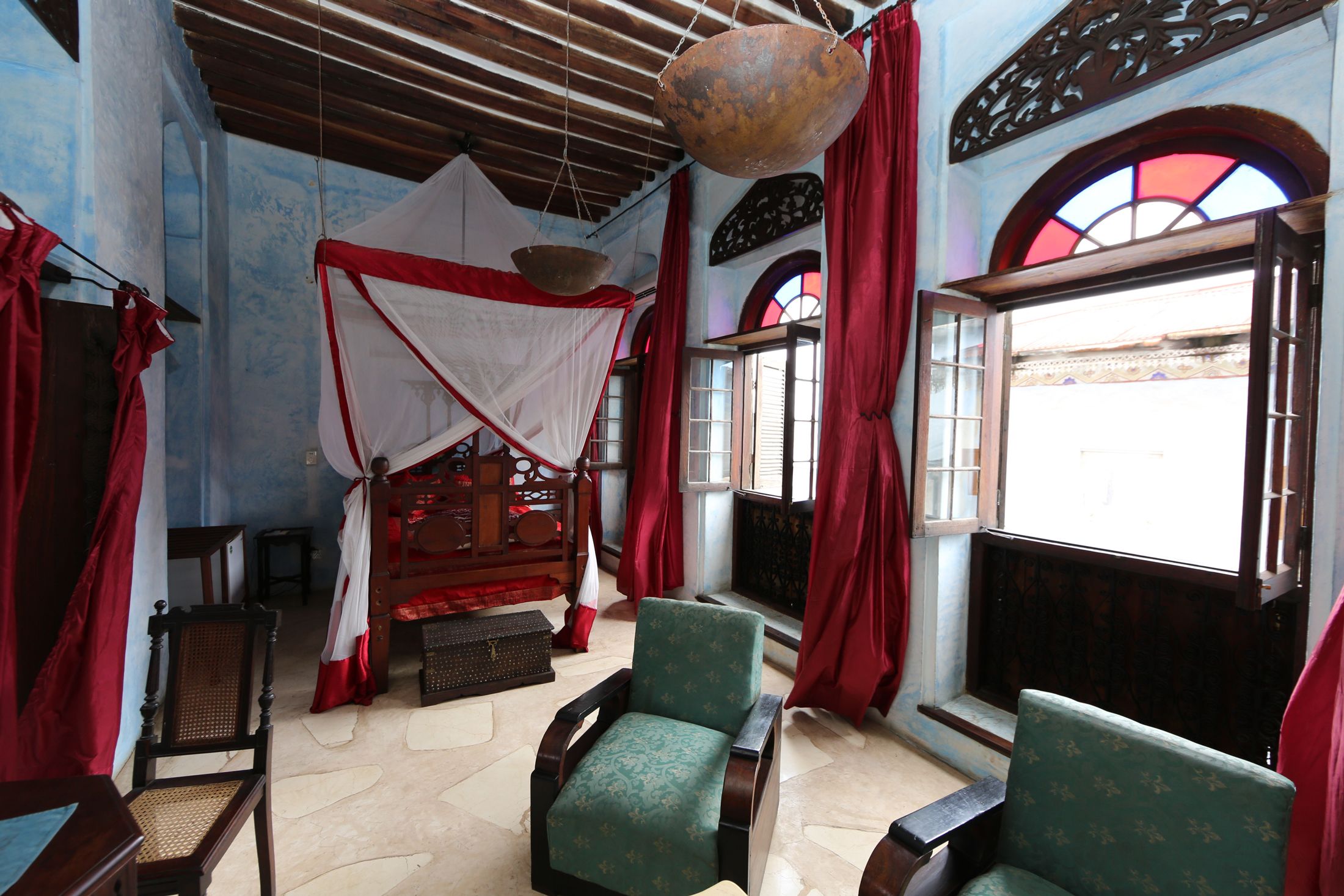 A room with a bed, two chairs, a hanging light fixture, and a window with red curtains.