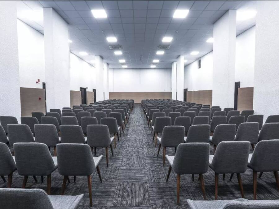 Una sala de conferències amb files de cadires grises, un sòl amb moqueta i parets blanques.