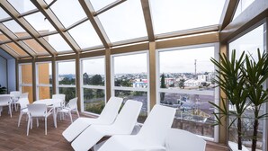 Una habitación iluminada por el sol con sillones y mesas blancos, y grandes ventanales con vistas a un paisaje urbano.