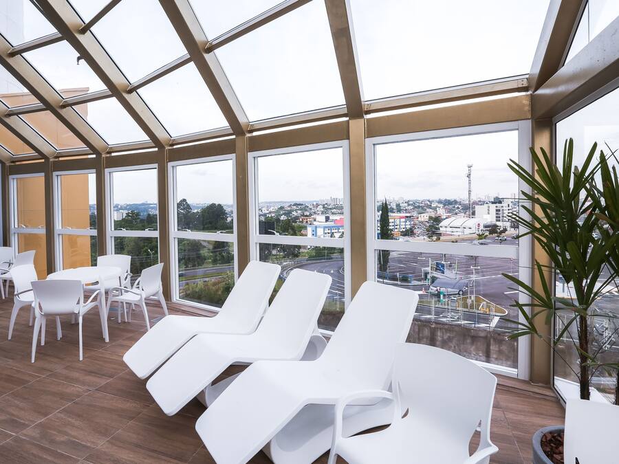 A sunlit room with white lounge chairs and tables, large windows offering a view of a cityscape.