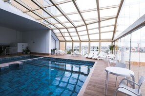 A modern indoor swimming pool with clear glass ceilings and a city view.