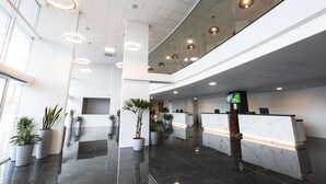 Un moderno vestíbulo de hotel con una recepción, plantas en macetas y una barandilla de vidrio que da al vestíbulo.