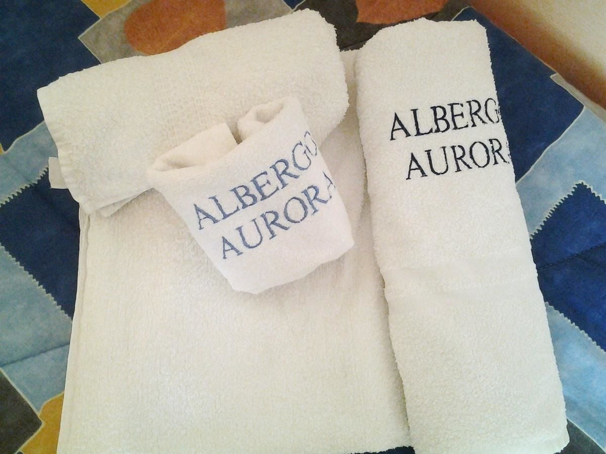 Deux serviettes blanches sur lesquelles est imprimé le nom « ALBERGO AUROR », posées sur un lit dont la couette présente un motif matelassé.