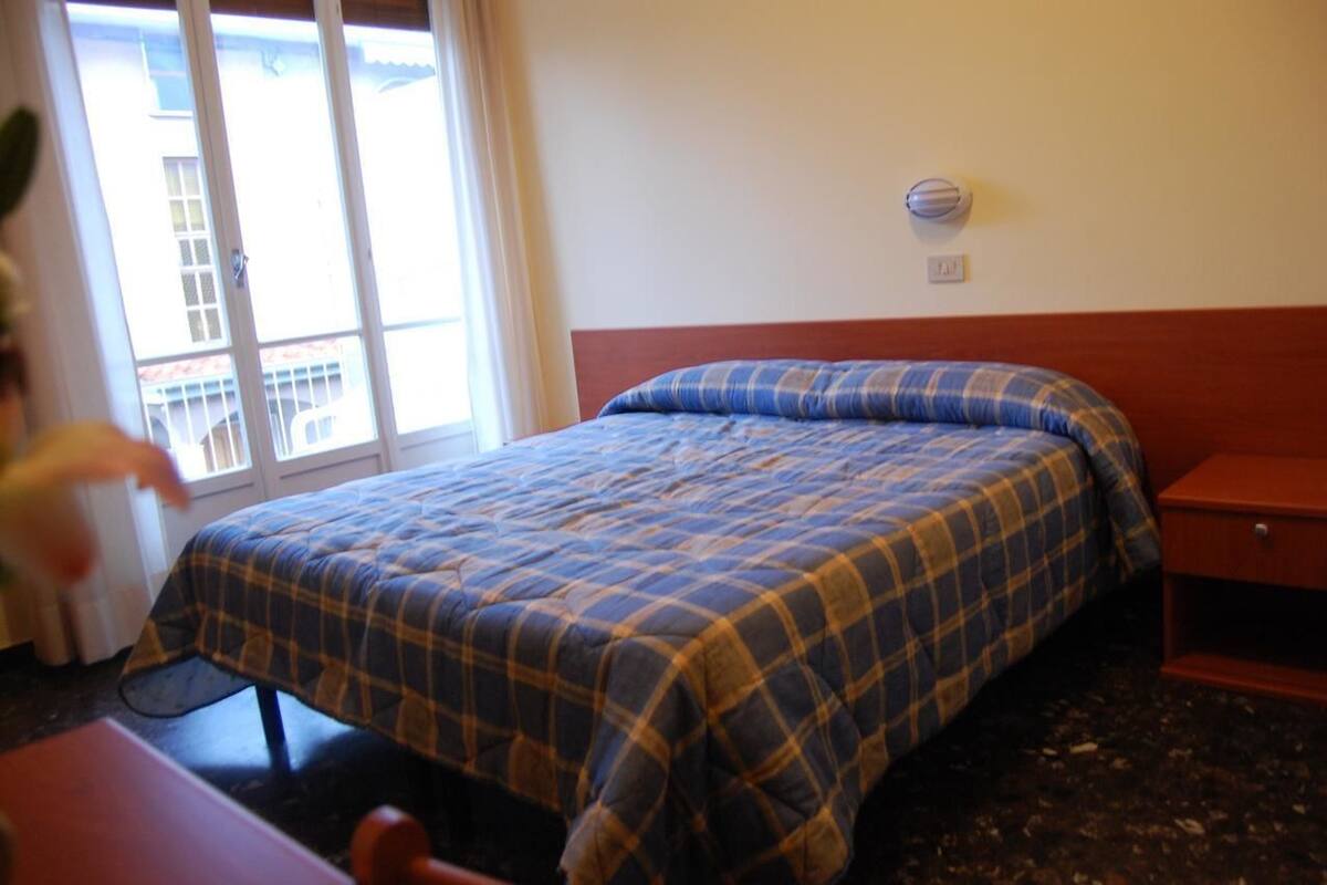 A hotel room with a bed, a nightstand, a window with a view of buildings, and a wall-mounted light fixture.