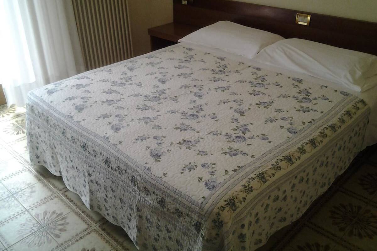 Ein Bett mit floralem Überwurf und Spitzenbordüre, ein hölzernes Kopfende sowie ein Nachttisch mit Spiegel.