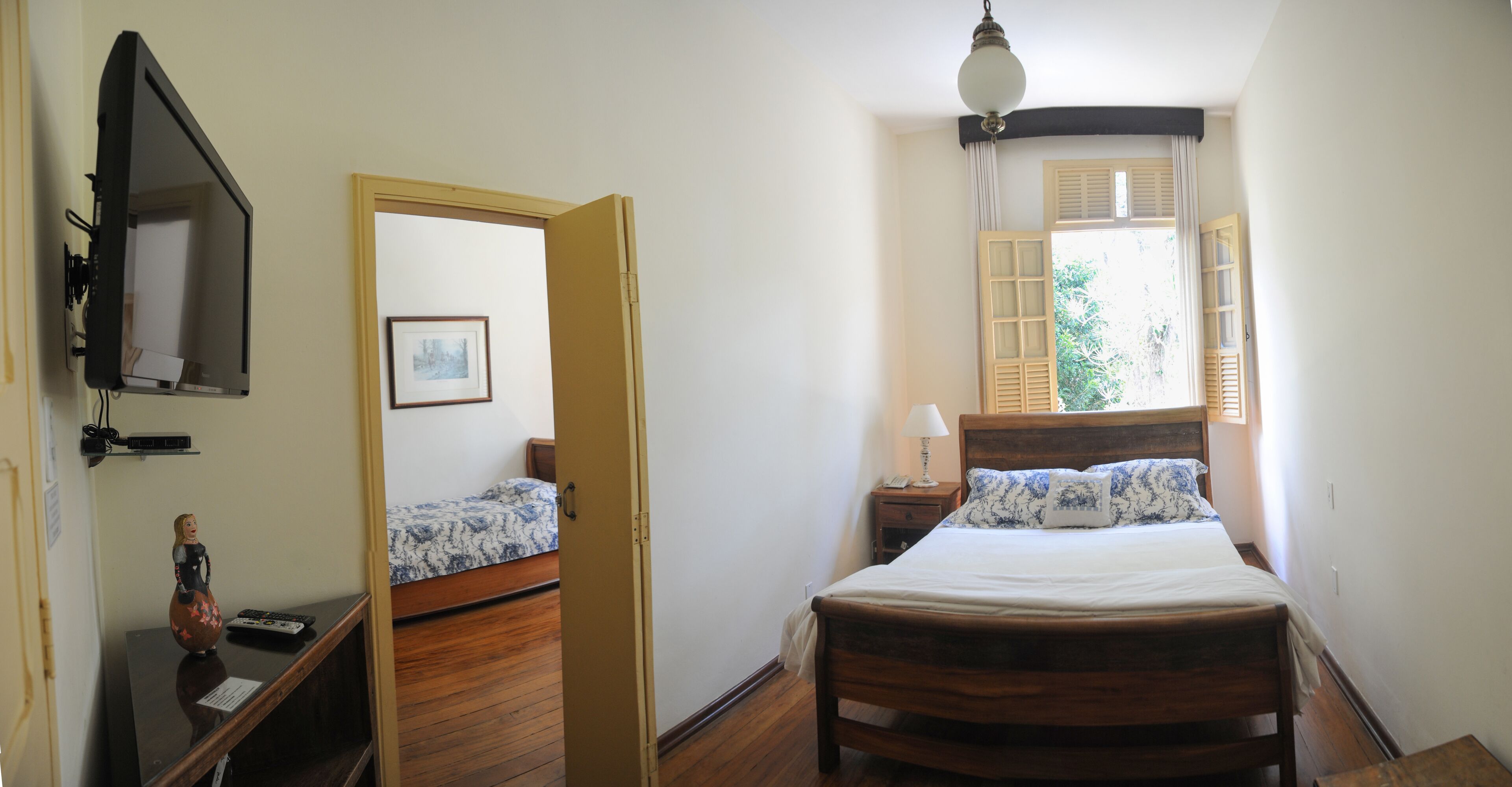 Une chambre à coucher avec un lit en bois, une table de chevet avec une lampe, un petit bureau avec un ordinateur et une fenêtre avec des rideaux.