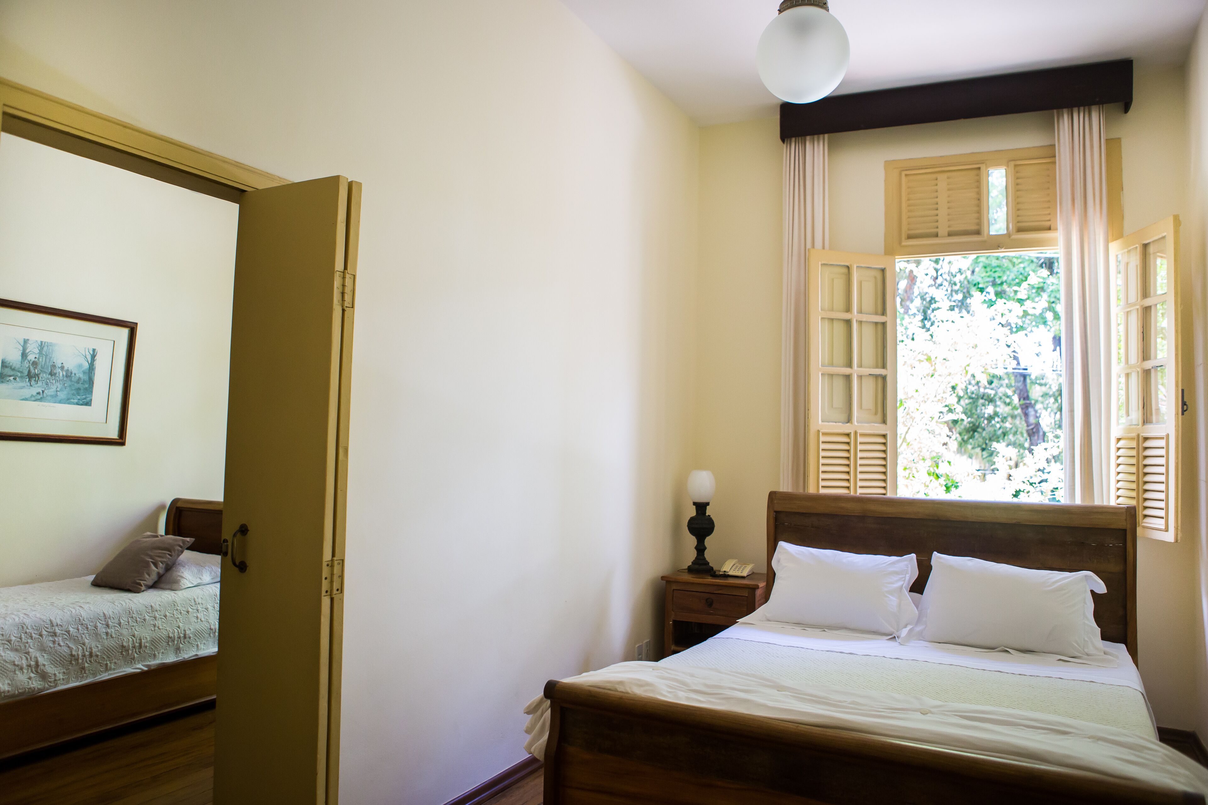 A bedroom with a wooden bed, a nightstand with a lamp, a window with shutters, and a door leading to another room.