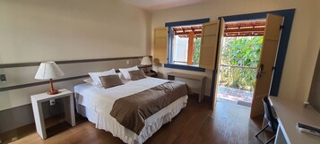 Quarto de hotel com cama, duas mesas de cabeceira, abajur, janela com persianas e vista para o verde lá fora.