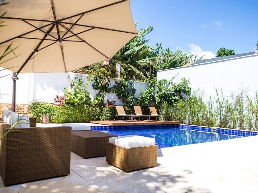 Un espace piscine aménagé avec des meubles en osier, un grand parasol et un mur blanc agrémenté de plantes.