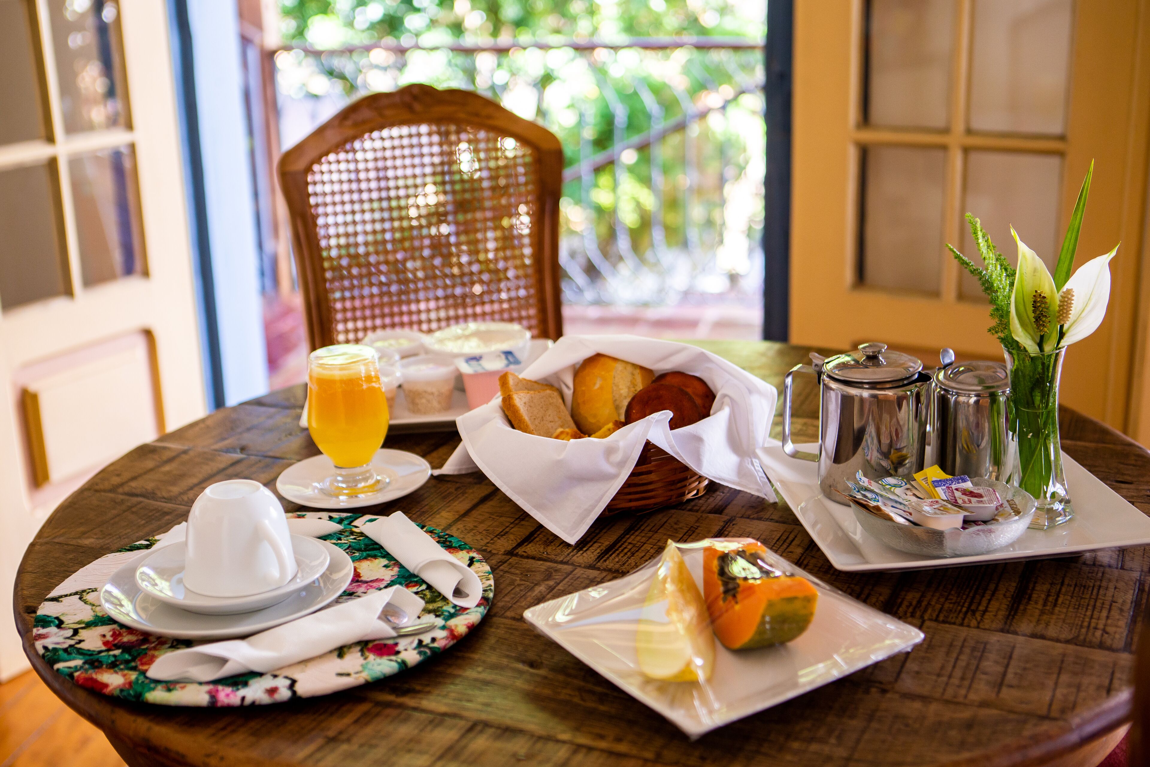 Una mesa de desayuno con platos, pan, un vaso de zumo de naranja y un florero con flores.