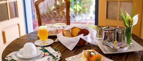 Una mesa de desayuno con platos, pan, un vaso de zumo de naranja y un florero con flores.