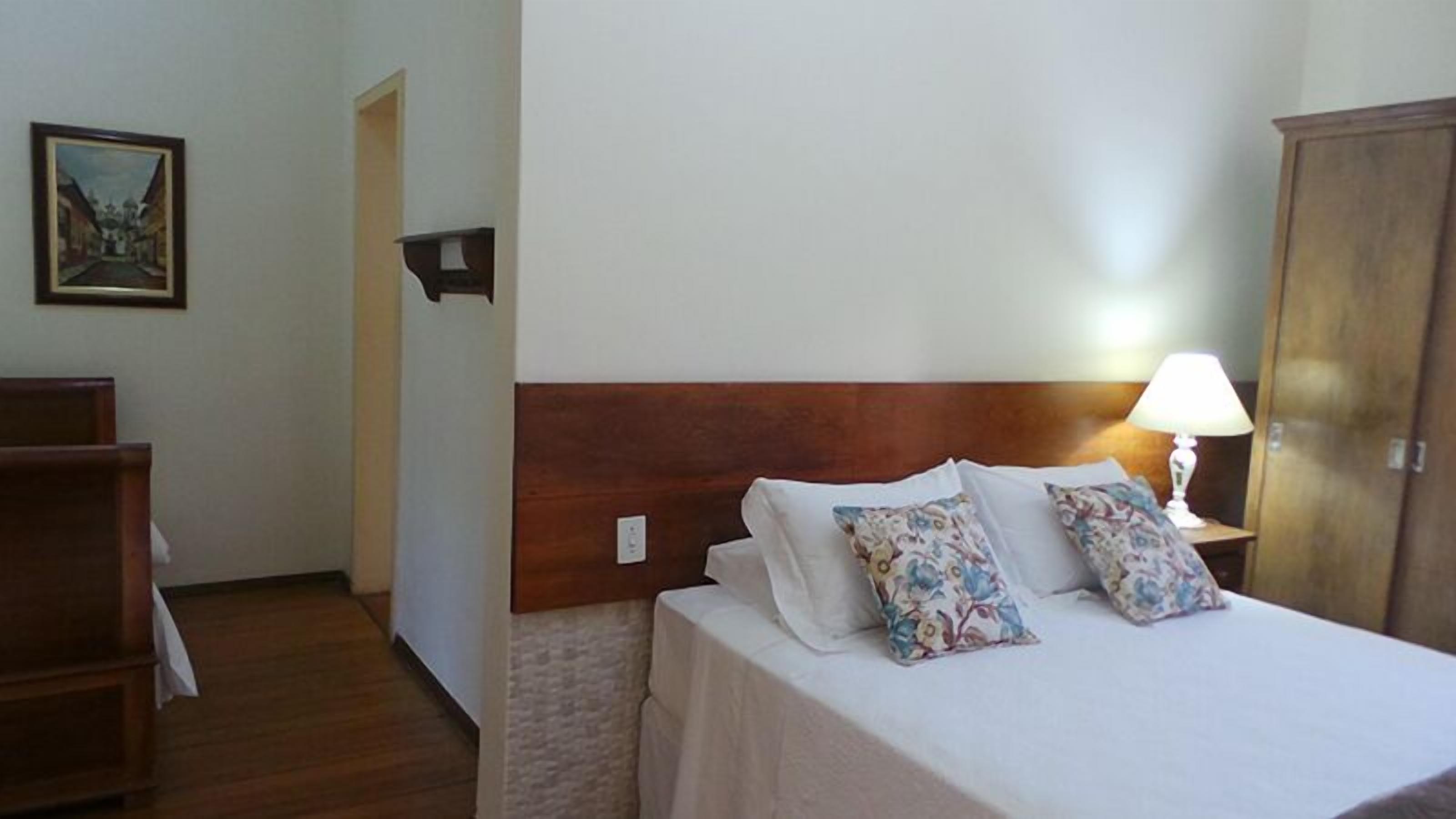 A hotel room with a wooden headboard, a bed with white linens and floral pillows, a nightstand with a lamp, and a painting on the wall.