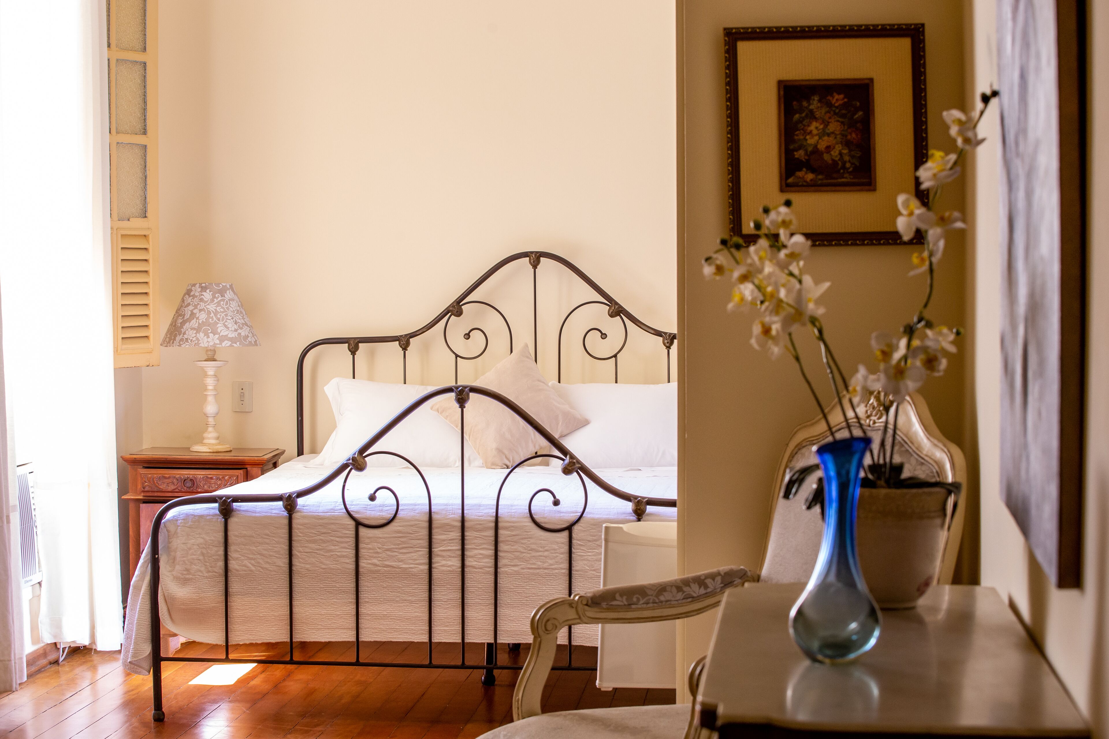 A bedroom with a metal bed frame, a bedside table with a lamp, a chair, and a vase with flowers.