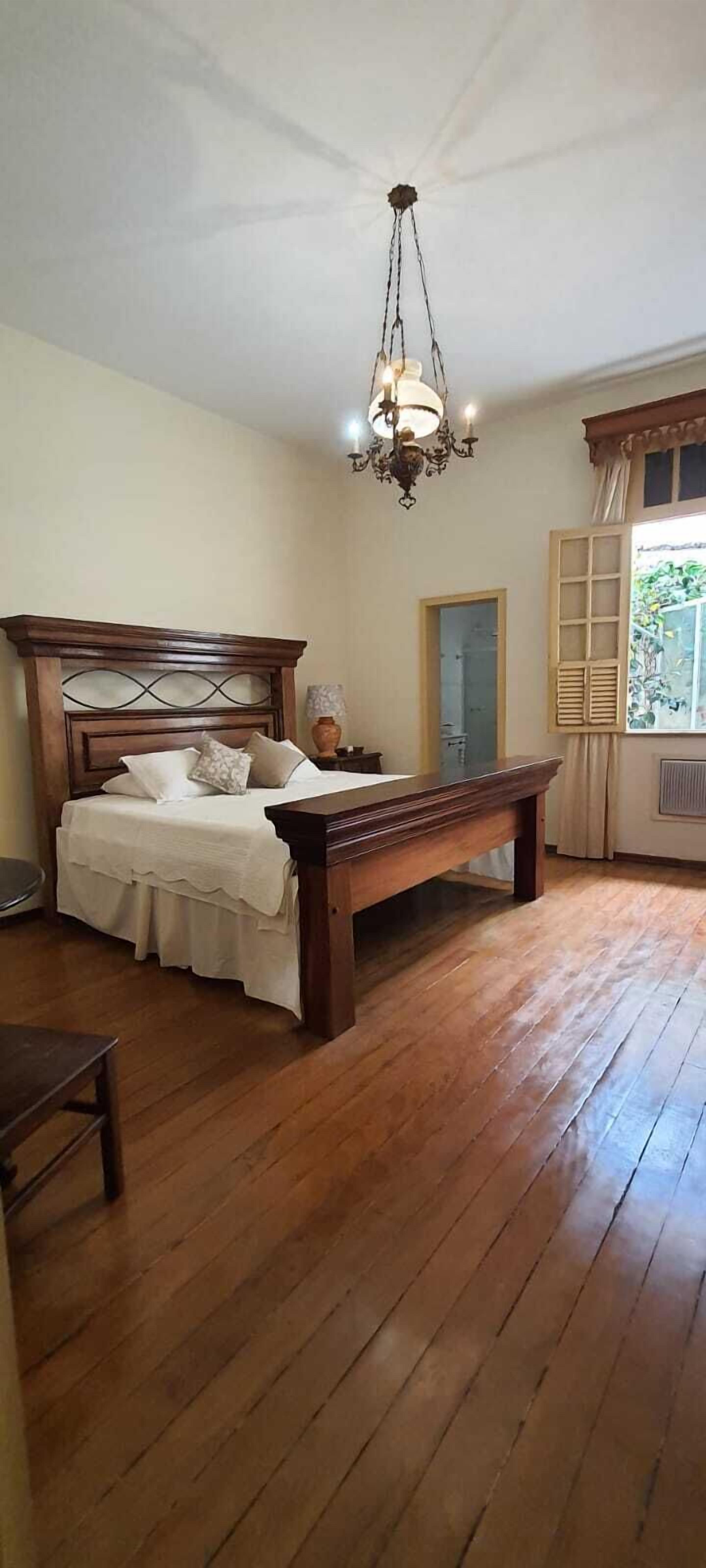 Un dormitori amb un llit de fusta, una panxa, una làmpada penjant i una finestra amb cortines.