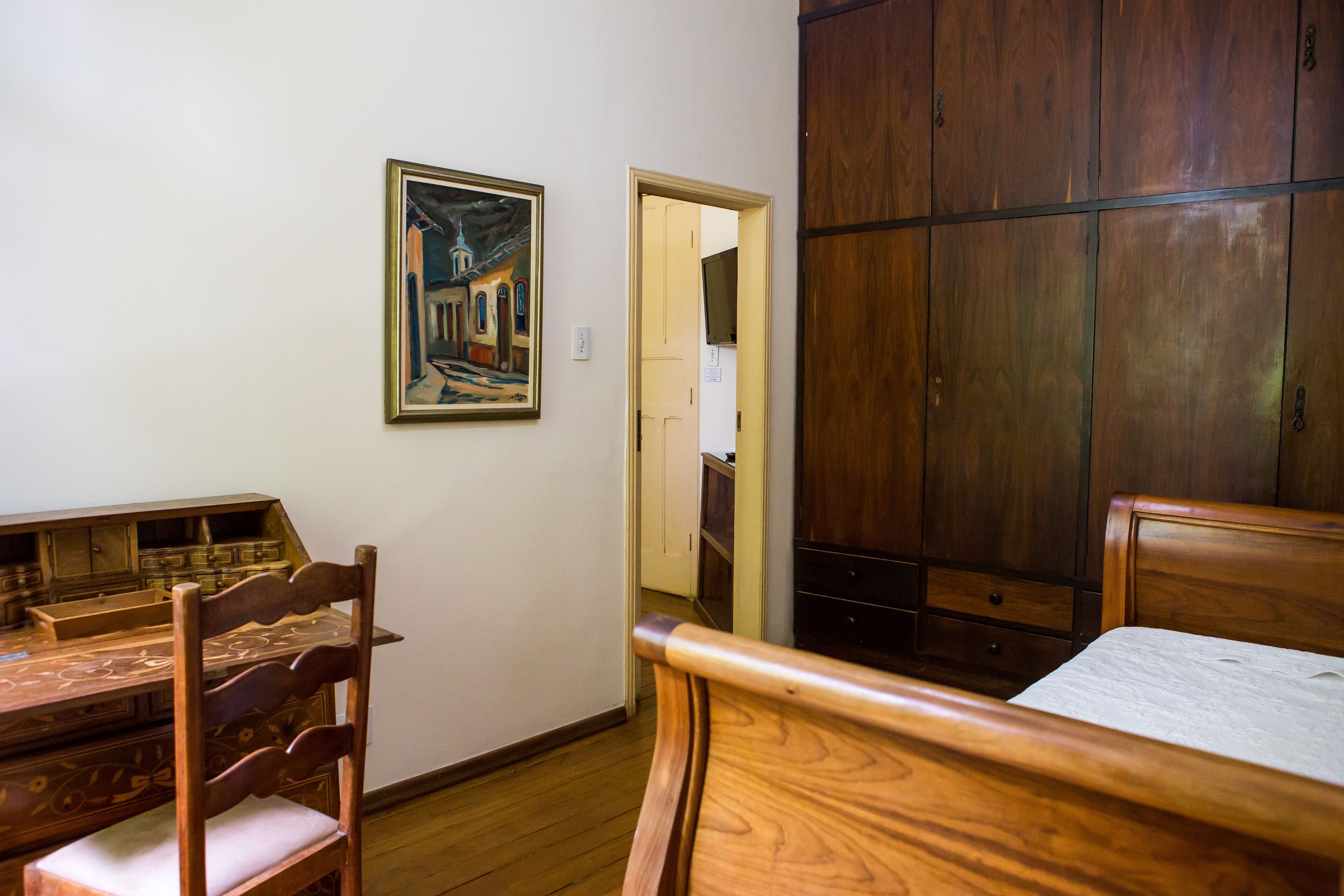 A room with a wooden bed, a wooden desk, and a chair. There is a painting on the wall and a wardrobe.