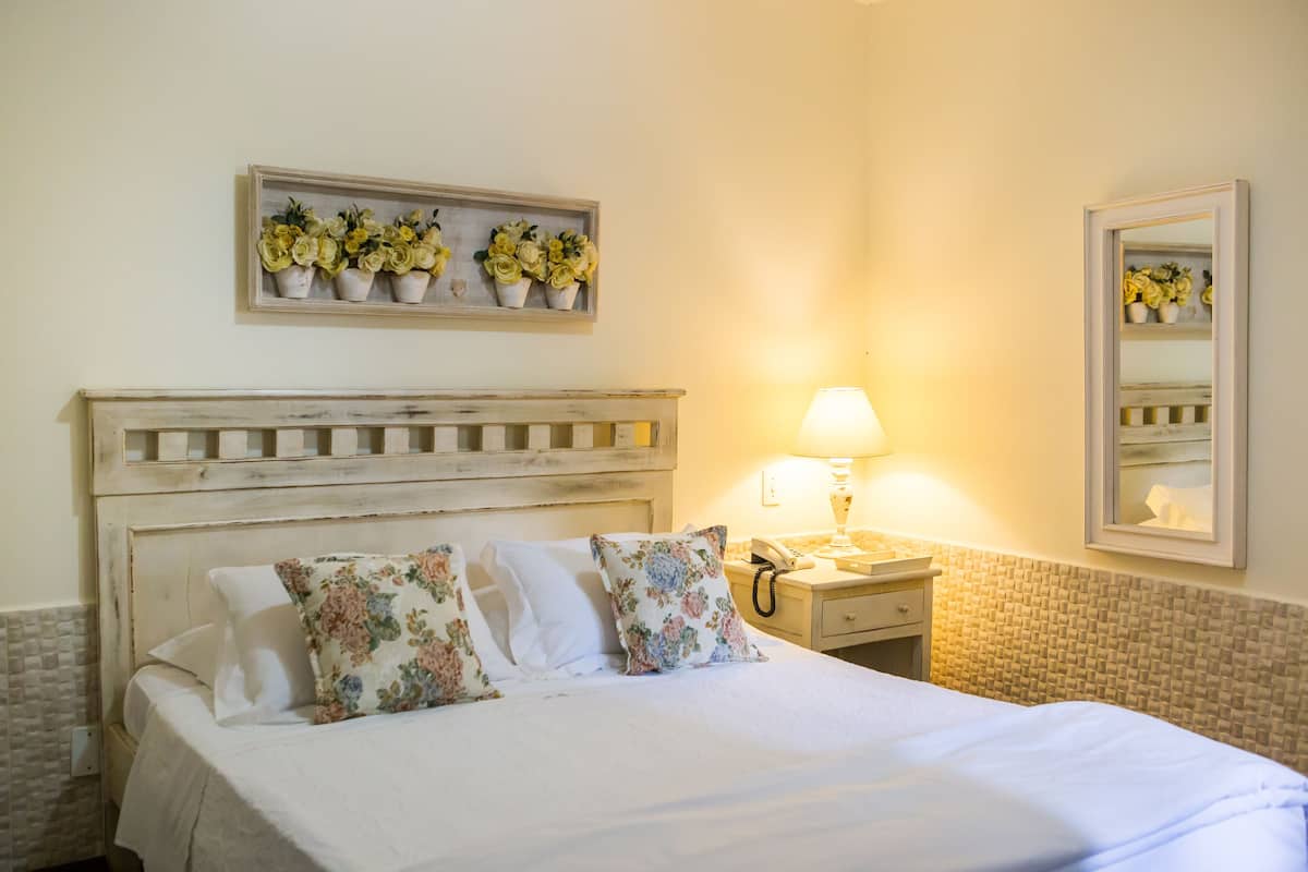 A neatly made bed with floral pillows, a bedside lamp, a small bedside table, and a mirror on the wall.