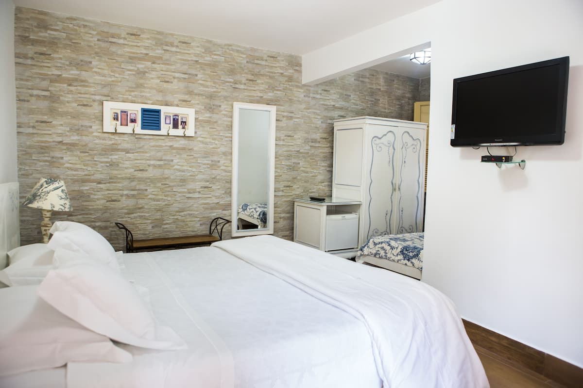 A bedroom with a stone-patterned wall, a bed with white bedding, a wardrobe, a mirror, and a flat-screen TV mounted on the wall.