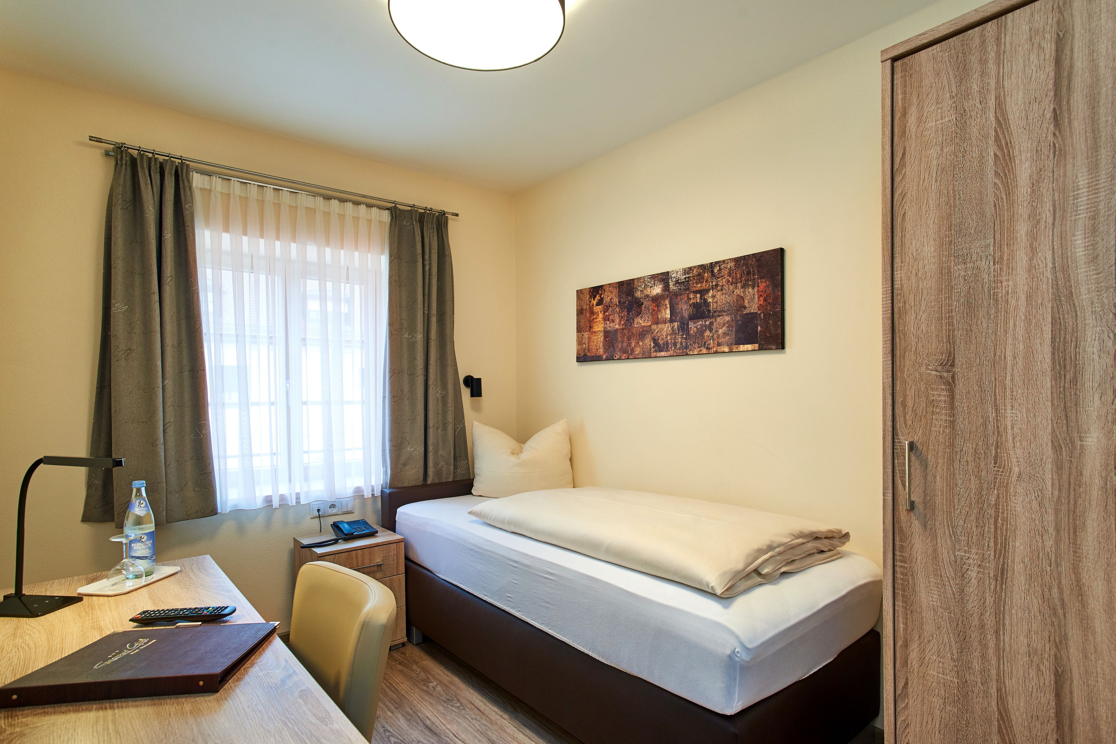 A hotel room with a bed, a desk with a lamp, a chair, a desk lamp, a water bottle, a remote control, and a painting on the wall.