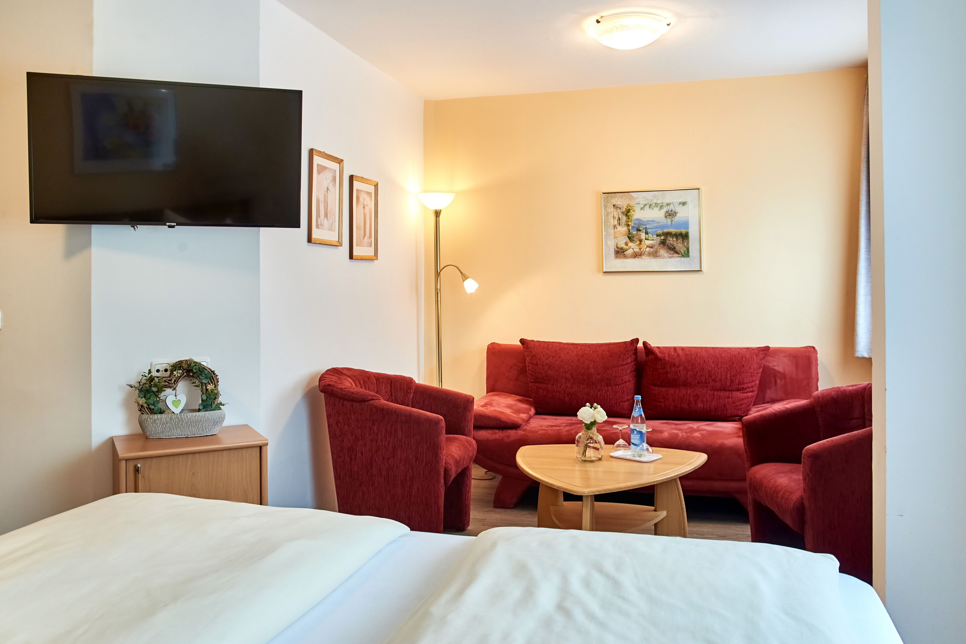 A hotel room with a bed, a red sofa, a small table with a vase of flowers, a lamp, and a painting on the wall.