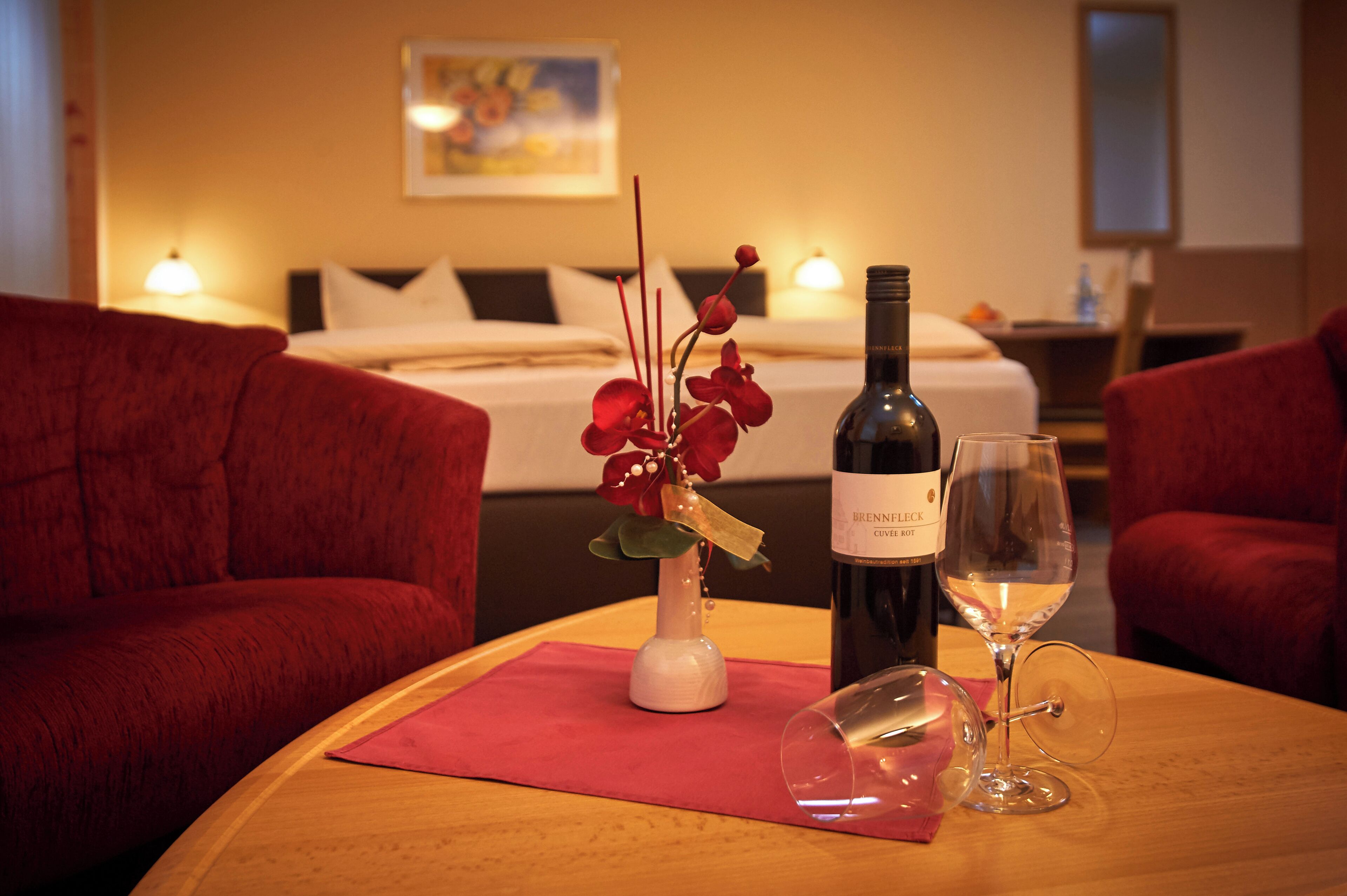 Ein Hotelzimmer mit einem Bett, roten Polstersesseln, einem Holztisch mit einer Weinflasche und Gläsern sowie einer Vase mit roten Blumen.