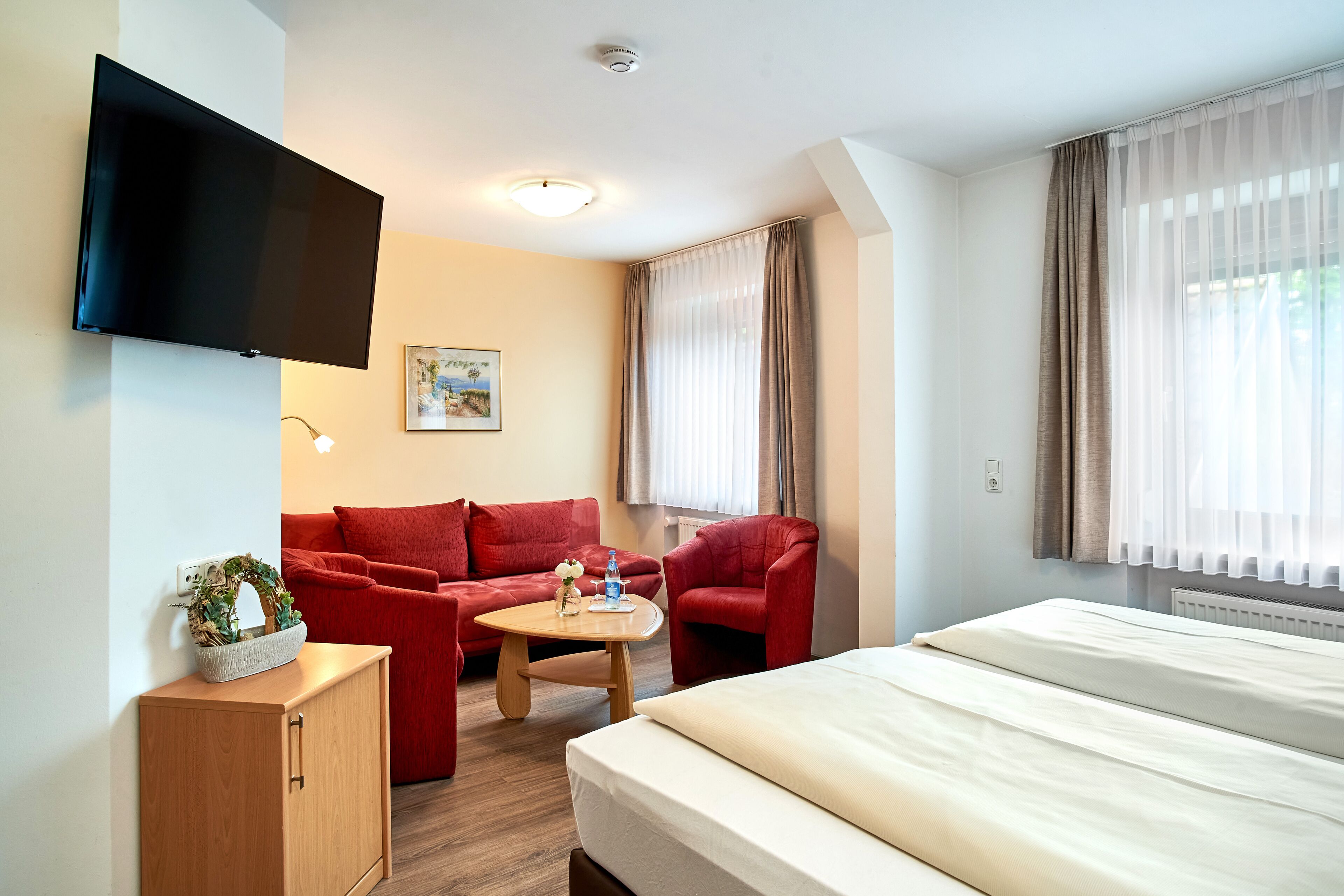 A hotel room with a bed, a red sofa, a red armchair, a small wooden table, a TV mounted on the wall, and a window with curtains.