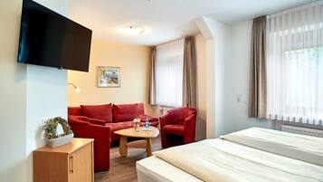 A hotel room with a bed, a red sofa, a red armchair, a small wooden table, a TV mounted on the wall, and a window with curtains.