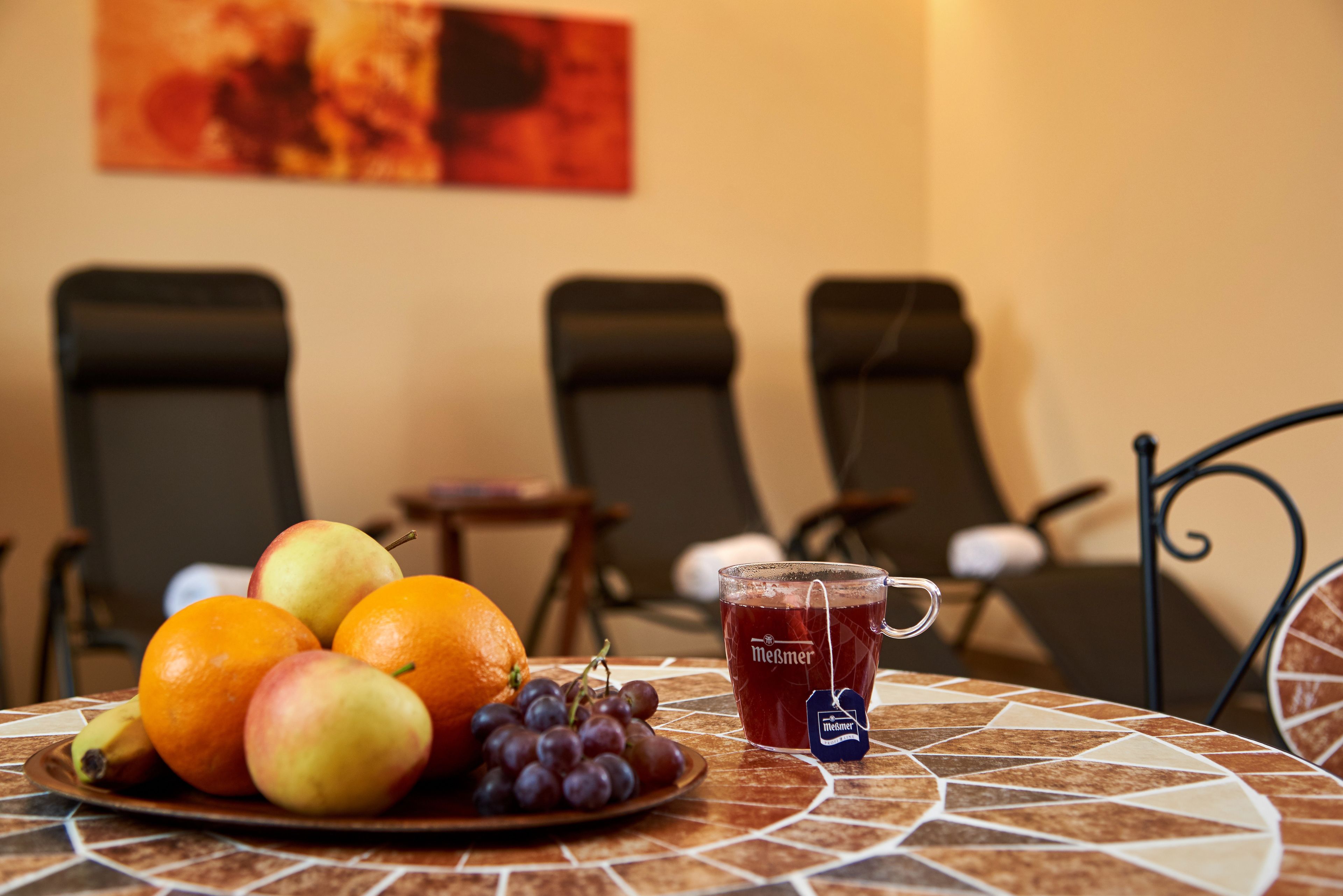 Una mesa con un plato de fruta y una taza, sillas al fondo y un cuadro en la pared.