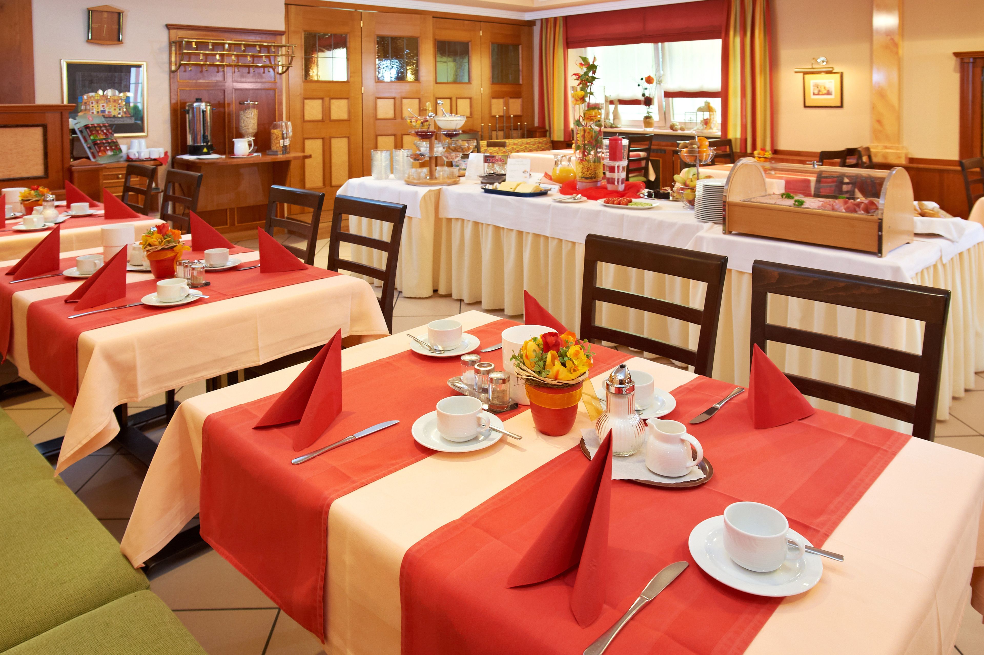 Un comedor de hotel con varias mesas dispuestas para el desayuno, cada una con servilletas rojas, platos blancos y una variedad de alimentos para el desayuno.