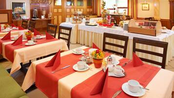 Un comedor de hotel con varias mesas dispuestas para el desayuno, cada una con servilletas rojas, platos blancos y una variedad de alimentos para el desayuno.
