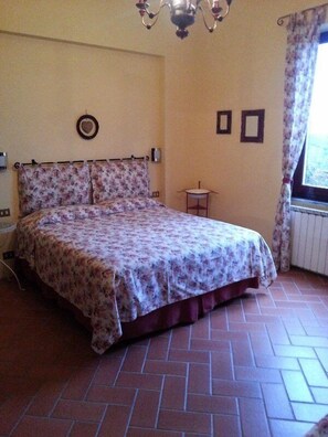 A bedroom with a floral bedspread, a chandelier, a small table, and a window with curtains. - Gli Aceri Bed and Breakfast (Arcola)