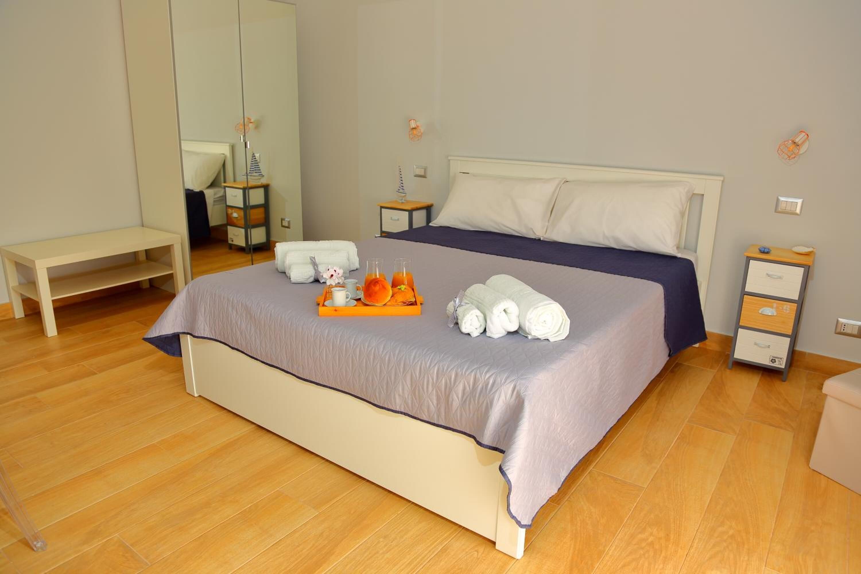 Uma cama bem arrumada com um edredom roxo, uma mesa de cabeceira com abajur e um pequeno banco de madeira.