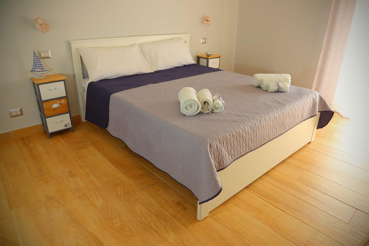 Una cama bien hecha con una colcha azul y blanca, una mesita de noche con una lámpara y una pequeña mesa con otra lámpara.