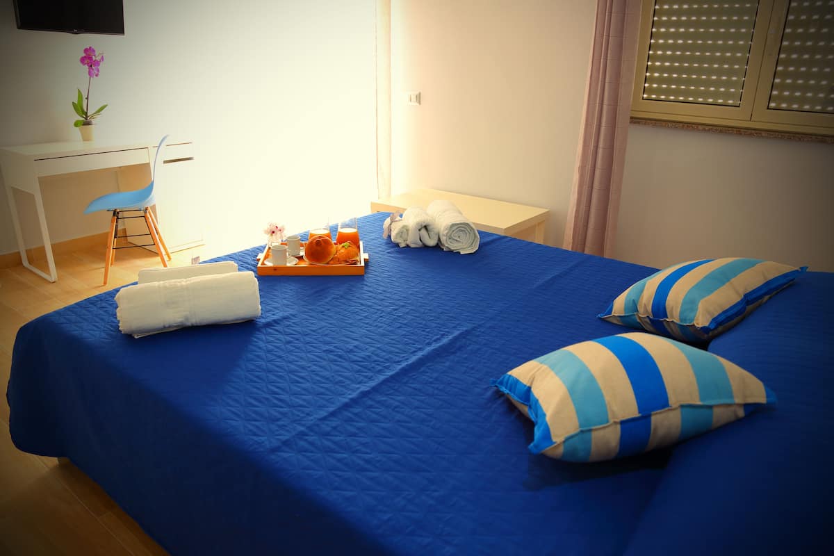 Una cama bien hecha con ropa de cama azul, dos almohadas a rayas y una bandeja con una toalla, zumo de naranja y una pequeña planta.