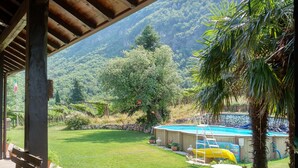A covered patio with a view of a backyard featuring a swimming pool, a wooden deck, and lush greenery. - Vallarom (Avio)