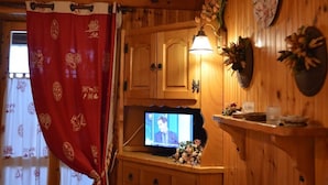 Een houten interieur met een televisie op een houten standaard, een klok aan de muur en een rood gordijn met decoratieve patronen.