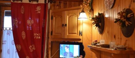 Un interior de madera con un televisor sobre un soporte de madera, un reloj en la pared y una cortina roja con motivos decorativos.