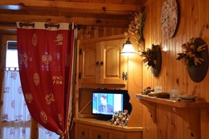 Un interior de madera con un televisor sobre un soporte de madera, un reloj en la pared y una cortina roja con motivos decorativos.