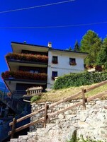 Un edificio de varios pisos con balcones decorados con flores rojas, una escalera de piedra y un cielo azul despejado.