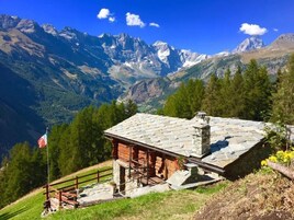 Una cabaña de madera con techo de piedra, rodeada por una valla de madera, con montañas y árboles al fondo.