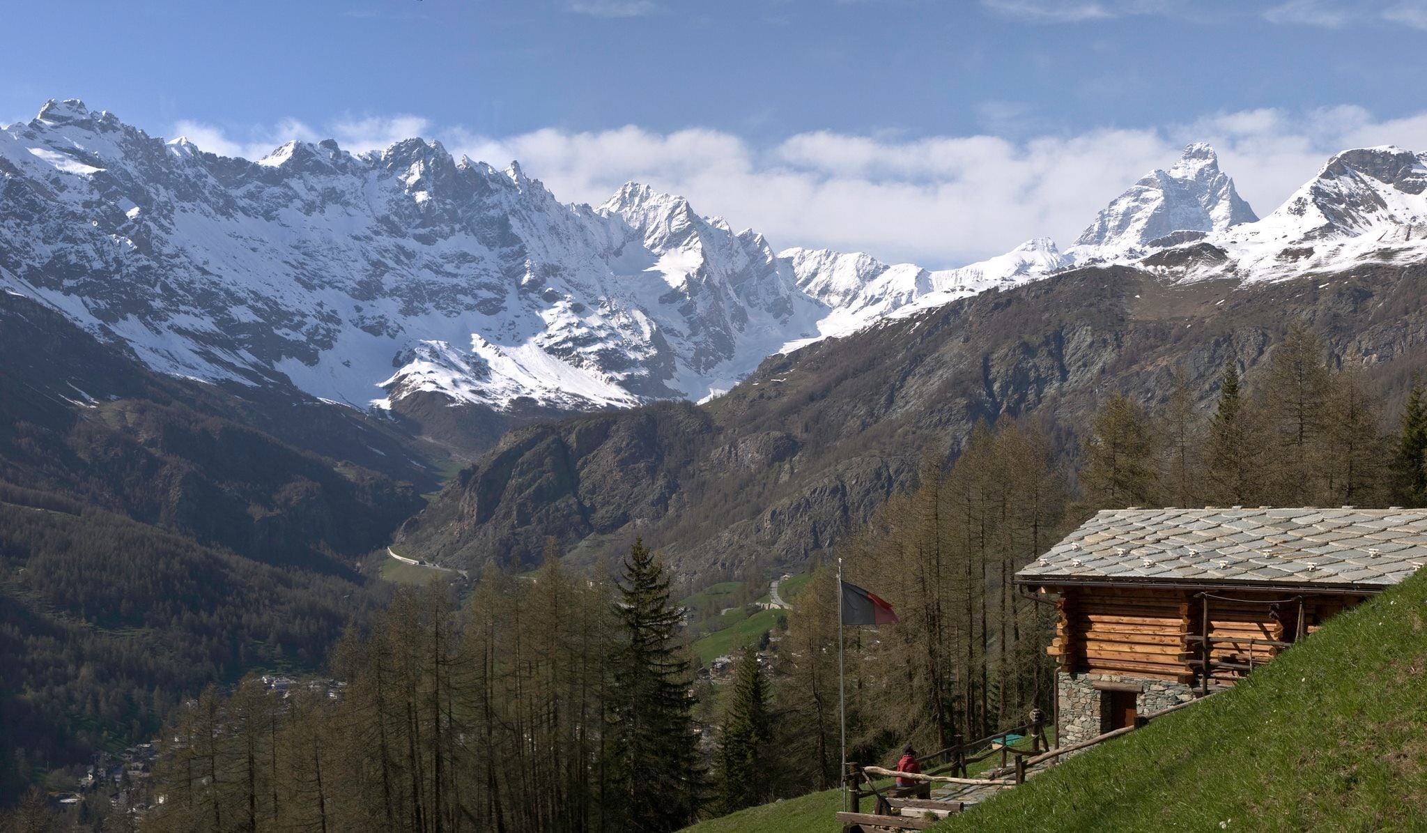A mountain lodge with a stone roof, surrounded by snow-capped peaks and forested valleys.