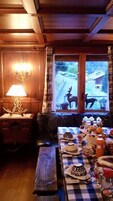 Una habitación acogedora con un banco de madera, una mesa dispuesta con varios platos, y una ventana que ofrece vistas a un ciervo y un edificio.