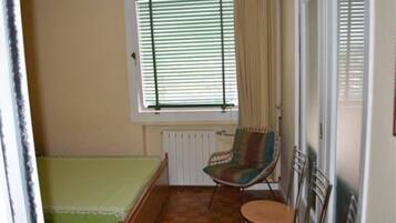 Ein kleines Zimmer mit einem Einzelbett, einem Sessel, einem Tisch und einem Fenster mit Jalousien.