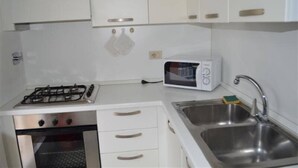 A compact kitchen with white cabinets, a gas stove, a microwave, and a stainless steel sink. - Residence Cristallo (Alassio)