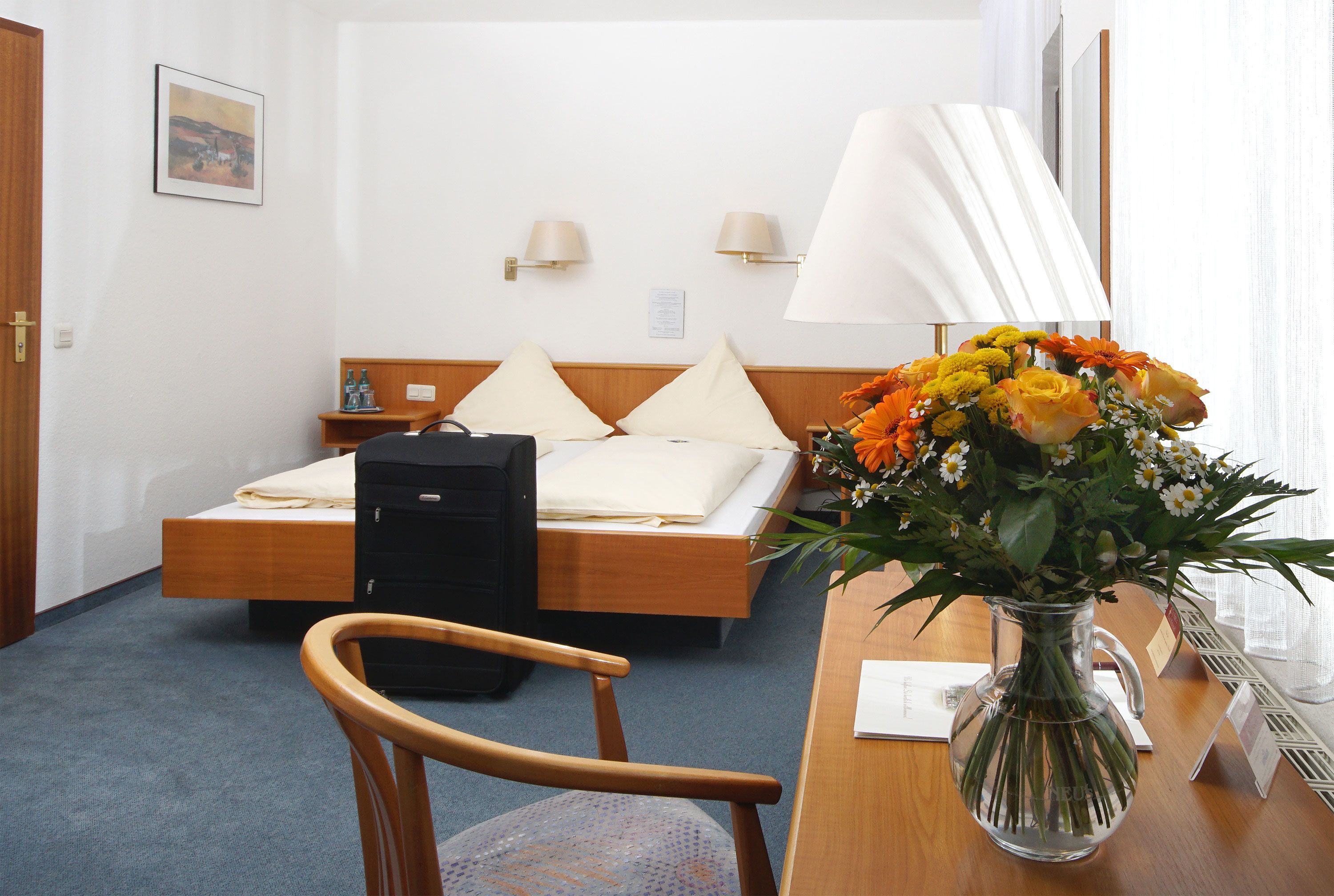 Une chambre d’hôtel comprenant un lit, des tables de chevet, des lampes, un bureau avec un vase de fleurs et une chaise.