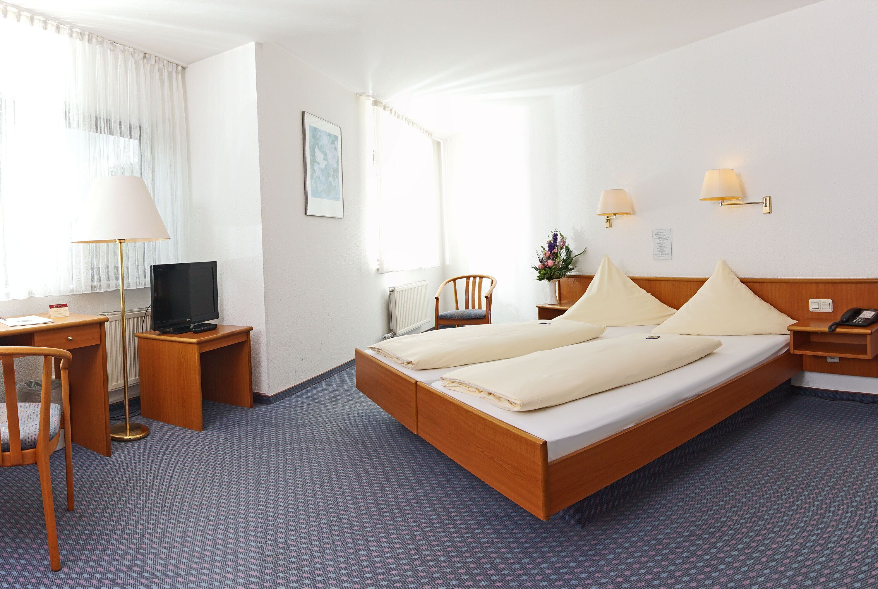 Ein Hotelzimmer mit einem hölzernen Bett, einem Schreibtisch mit Stuhl, einem Fernseher und zwei Nachttischlampen.