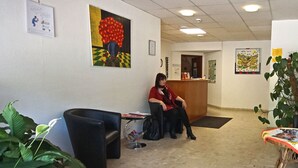 Een receptie met een zittende receptionist, een zwarte bank, een klein tafeltje met tijdschriften, en een kamerplant.